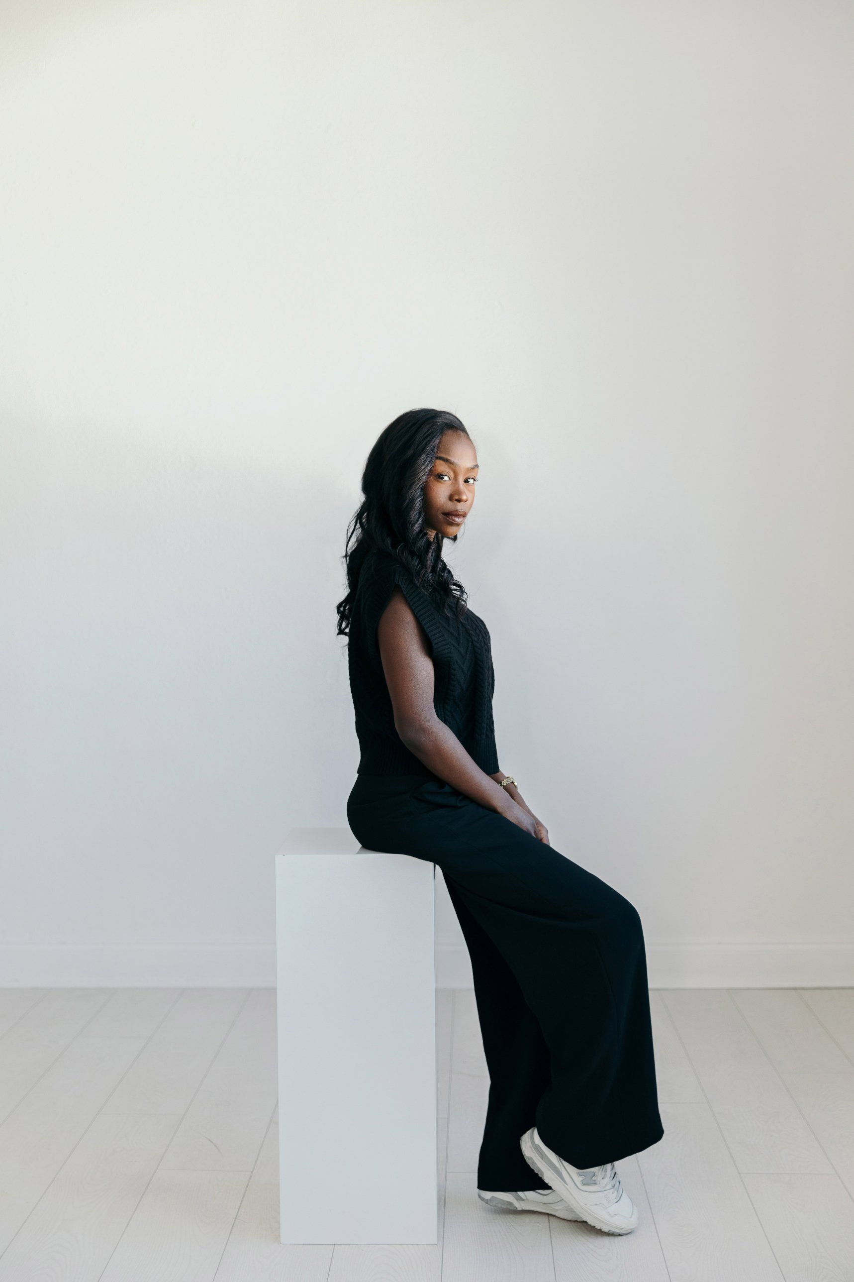 Avrielle Muse seated on a white pedestal in a minimal studio, looking toward the camera